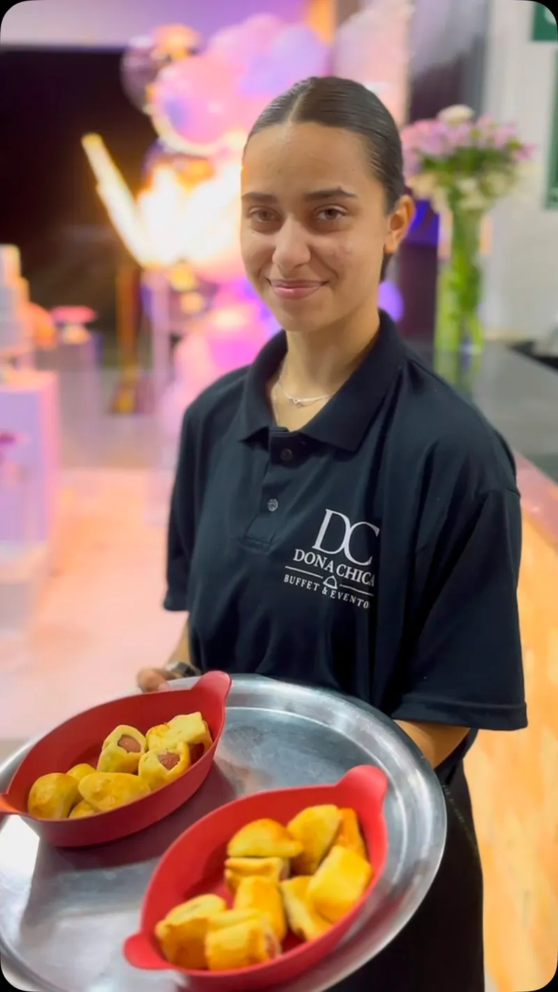 Integrante da equipe Dona Chica, de polo preta com o monograma DC, segurando bandeja de salgados